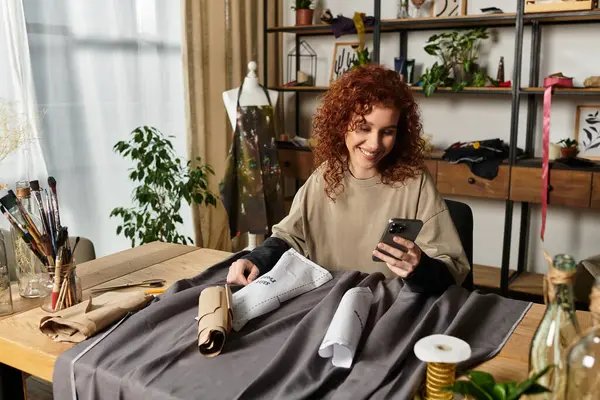 Şık bir çalışma alanındaki kumaş ve aletlerin arasında telefonundaki neşeli zanaatkar kadın..