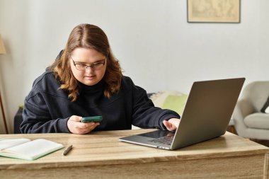 Down sendromlu kendine güvenen genç bir kadın dizüstü bilgisayarı ve telefonu ile evde oturuyor..