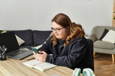 Down sendromlu genç bir kadın ev masasında telefonunu kullanırken gülümsüyor..