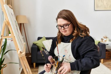 Down sendromlu genç bir kadın evde resim yapıyor. Etrafı sanat malzemeleriyle çevrili..