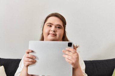 Down sendromlu genç bir kadın evinde rahatça oturup, kendinden emin bir şekilde tablet tutuyor..
