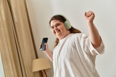 Down sendromlu genç bir kadın evinde neşeyle dans ediyor, rahat kıyafetler ve kulaklıklar takıyor..