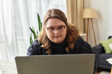 Parlak ve havadar ev ortamında dizüstü bilgisayarına odaklanmış Down sendromlu genç bir kadın görülüyor..