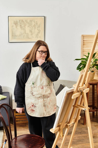 Молодая женщина с синдромом Дауна рассматривает свои работы, стоя в уютной домашней студии.