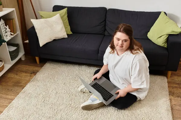 Down sendromlu neşeli genç bir kadın rahat bir odada dizüstü bilgisayarına odaklanmış bir şekilde yerde oturuyor..