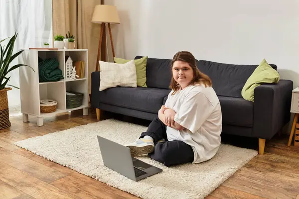 Down sendromlu genç bir kadın rahat bir odada dizüstü bilgisayarını kullanarak yerde oturuyor..