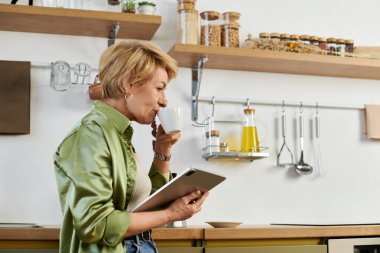 Son sınıf öğrencisi kadın kahvesini yudumluyor, güneşli bir mutfakta tabletine hayran, sabah rutininin tadını çıkarıyor..