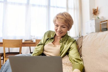 Üst düzey bir kadın sıcak ve davetkar bir yaşam alanında laptopunu kullanırken parlak bir şekilde gülümsüyor..