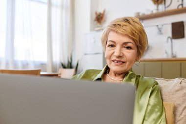 Parlak bir oturma odasında internetteyken kısa sarı saçlı yaşlı kadın gülümsüyor..