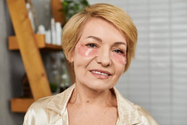 Güzel, son sınıf öğrencisi bir kadın gözlerinin altına nemlendirici bantlar yapıştırıyor, neşe ve kendine güvenle parlıyor..