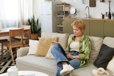 Keyifli, son sınıf öğrencisi bir kadın, rahat oturma odasında dinlenirken tabletini okuyor..