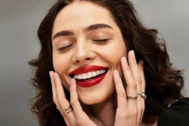 Genç bir kadın elleri yanaklarında gülümsüyor, diş tellerini ve kalın kırmızı dudaklarını gösteriyor..