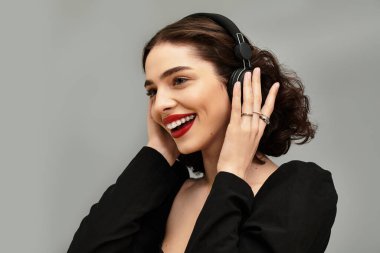 Telleri ve kırmızı dudakları olan genç bir kadın kulaklıklarıyla müzik dinlerken gülümsüyor..