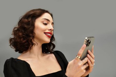 Telleri ve kırmızı dudakları olan neşeli genç bir kadın gri arka planda telefonuna gülümsüyor..