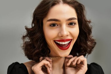 Genç bir kadın diş telleri ve koyu kırmızı dudaklarıyla gri bir zemine gülümsüyor..