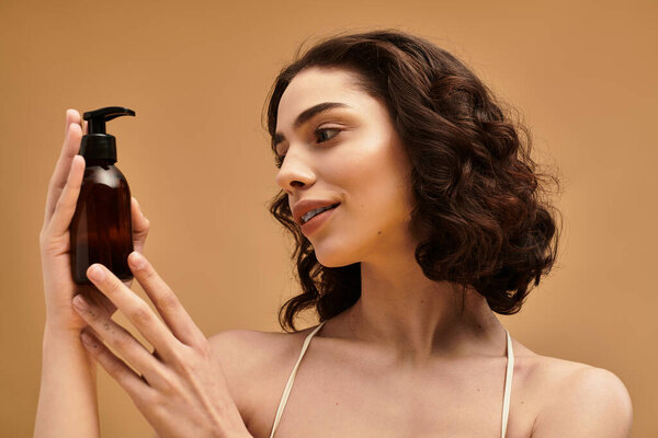 Молодая женщина подчеркивает свою рутину по уходу за кожей, улыбаясь и глядя на средство для очищения лица в теплой студийной обстановке.