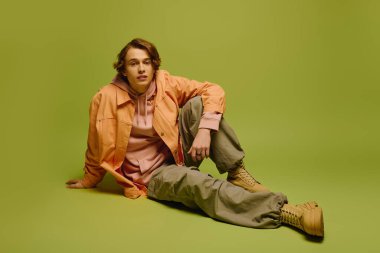 Şık bir genç adam, parlak yeşil bir arka fona rahatça oturur ve hiç çaba harcamadan serinkanlılığını yayar..