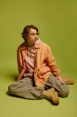 Modaya uygun genç bir adam, parlak yeşil bir zemine karşı gelişigüzel oturuyor ve modaya uygun tarzını sergiliyor..