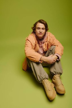 Moda kıyafetleri içinde genç bir adam canlı yeşil bir arka plana karşı gelişigüzel poz veriyor..