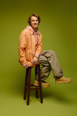 Şık bir genç adam bir taburede oturur, parlak bir arka plana karşı kıyafetini sergiler..