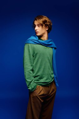 Moda kıyafetleri içindeki genç adam cesur mavi bir arka plana karşı kolayca poz veriyor, havalı bir tarzı ve zarafeti var..