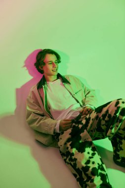 Moda kıyafetleri giymiş genç bir adam renkli bir arkaplanda poz verirken cazibe saçıyor..