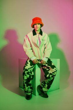 Genç bir adam eşsiz moda tarzını renkli bir ışıkla yıkanmış bir küpün üzerinde otururken gösteriyor..
