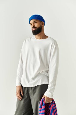 Beyaz gömlekli ve mavi bereli yakışıklı bir adam elinde çizgili bir eşyayla kendinden emin bir şekilde ayakta duruyor..