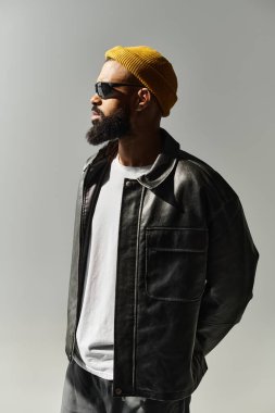 Siyah deri ceketli ve sarı bereli genç bir adam, açık bir arkaplanda güvenle poz veriyor..