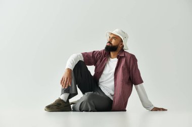 Şık bir adam, rahat bir şekilde yerde oturuyor, kendine güvenen bir tavır sergiliyor ve modern bir moda sergiliyor.