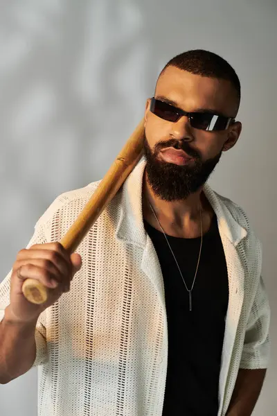 Sakallı ve güneş gözlüklü yakışıklı genç adam şık bir ortamda tahta bir yarasayla poz veriyor..