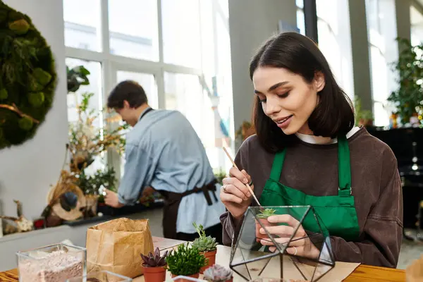 Genç bir sanatçı geometrik bir teraryumda küçük bir bitki düzenlerken, bir diğeri yakınlarda çalışır..