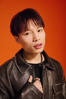 Genç bir Asyalı, parlak turuncu bir arka plana karşı deri ceketiyle cazibesini sergiliyor..