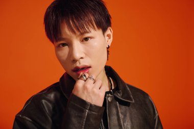 Genç adam, modaya uygun siyah deri bir ceketle turuncu bir arka planda tarz ve karizma sergiliyor..