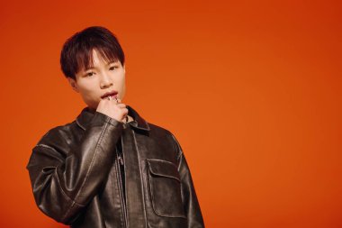 Siyah deri ceketli çekici bir genç adam parlak turuncu bir fonda duruyor..