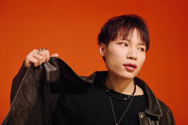 Deri ceketli şık, genç bir Asyalı turuncu arka planda rahatça poz veriyor..