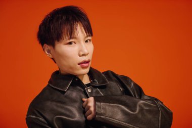 Kendine güveni olan genç bir adam, çarpıcı turuncu bir arka plana karşı şık bir dış giyim içinde poz veriyor..