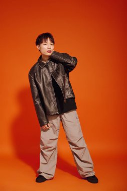 Deri ceketli ve rahat pantolonlu şık bir genç cesur turuncu arka planda poz veriyor..