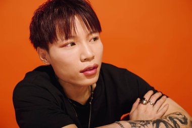 Şık dövmeleri olan Asyalı genç bir adam dalgın görünüyor. Parlak bir arka plana yaslanıyor..