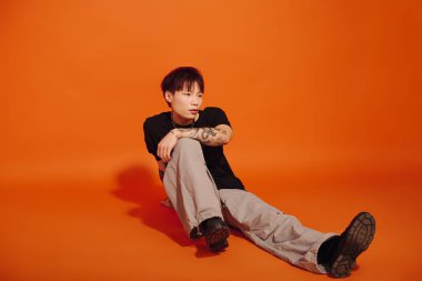 Şık bir Asyalı genç adam, cesur bir turuncu zemine karşı güven içinde yerde oturuyor..