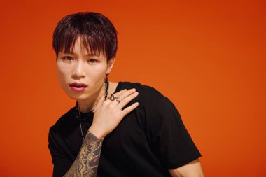 Şık bir Asyalı genç adam, cesur turuncu bir arka plana karşı moda ve dövmelerini sergiliyor..