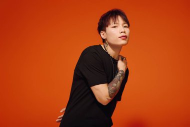 Şık bir Asyalı, canlı turuncu bir arka plana karşı, dövmelerini göstererek güvenle poz verir..