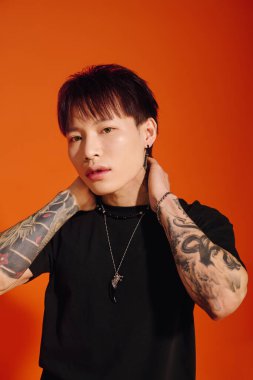 Kendine güvenen genç bir adam dövmelerini ve şık mücevherlerini canlı turuncu bir arka planda sergiliyor..