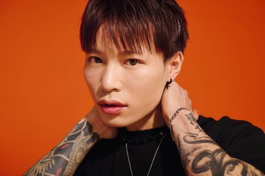 Yakışıklı Asyalı şık saçlı ve dövmeli adam turuncu bir duvara karşı güvenle poz veriyor..