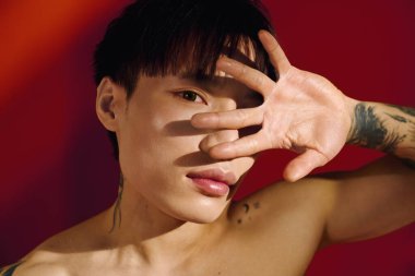 Yakışıklı Asyalı genç adam dövmelerini gösterirken bir eliyle yüzünü saklar..