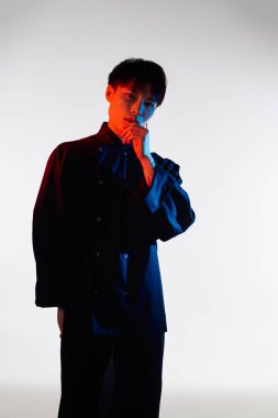 Genç bir Asyalı adam şık kıyafetlerini ve canlı renklerini göstererek düşünceli bir şekilde ayakta duruyor..