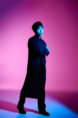 Yakışıklı Asyalı bir adam kollarını çarpıcı bir ışıkta kavuşturarak güvenle ayakta duruyor..