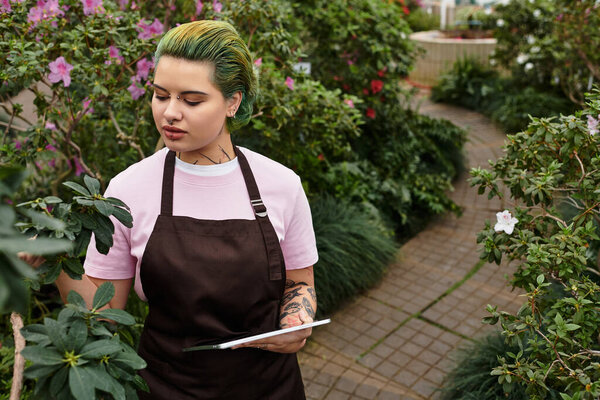 A young woman in an apron carefully examines plants in a colorful greenhouse surrounded by flowers.