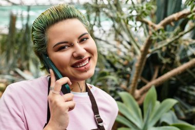 Serada gülümseyen genç bir kadın telefonuyla konuşuyor. Etrafı yeşilliklerle çevrili..