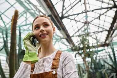 Genç bir kadın telefonuyla konuşuyor. Parlak, yeşil bir serada..